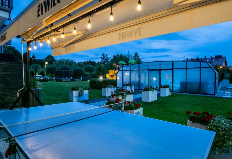 Outdoor hotel terrace at dusk with string lights, table tennis, glass-enclosed pool, lawn planters and playground