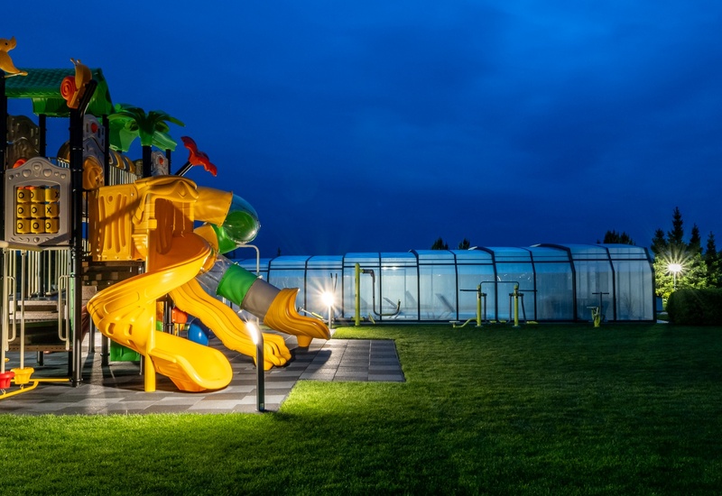 Illuminated children's playground with yellow slides on a manicured lawn beside a covered hotel pool at dusk.