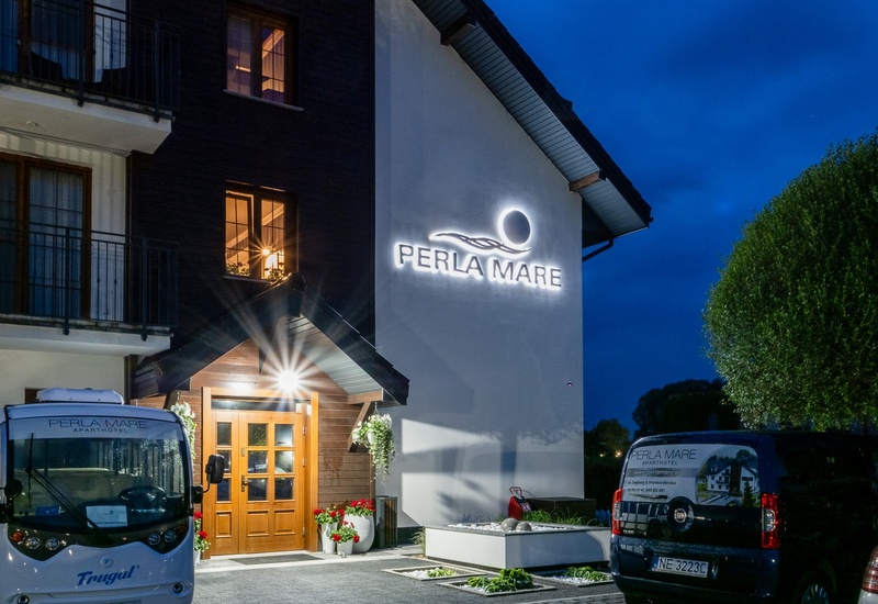 Illuminated Perla Mare aparthotel façade at night, warm wooden entrance, balconies, branded shuttle and parking, welcoming evening ambiance