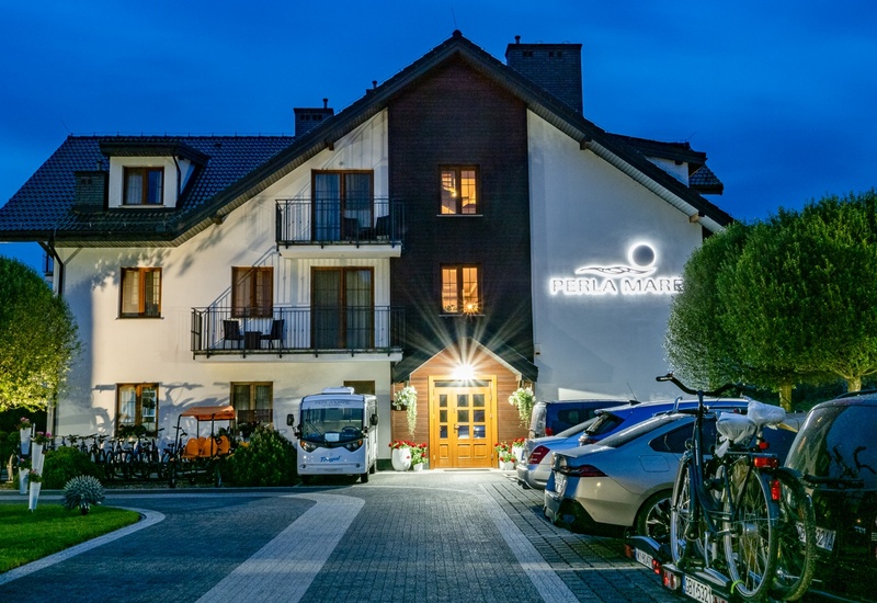 Three-story coastal hotel at dusk with a lit wooden entrance, balconies, illuminated "Perla Mare" sign, parked cars and bicycles