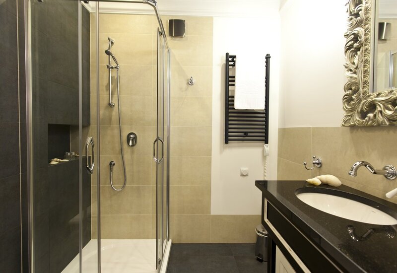 Hotel bathroom with glass shower and handheld fixture, toiletry niche, heated towel rack, black stone vanity and ornate mirror.