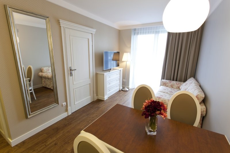 Bright guest suite with dining table and vase of flowers, floral sofa, TV on dresser, floor lamp, full-length mirror and balcony door with sheer curtains.