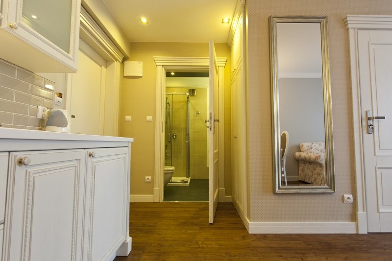 Small suite with kitchenette, hardwood floors, full-length mirror and en-suite bathroom with glass shower; floral sofa visible.