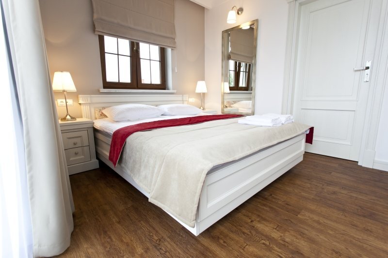 Bright double room with king bed, white wooden furnishings, bedside lamps, red bed runner, folded towels and hardwood floor.