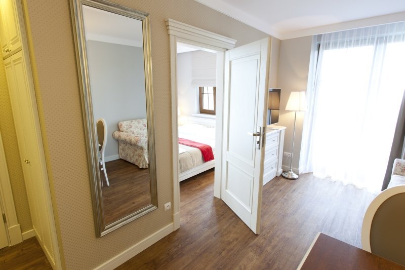 Bright suite with separate bedroom, king bed with red runner, floral sofa, full-length mirror, dresser, wooden floors and light-filled balcony.