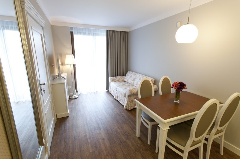 Bright hotel living area with floral sofa, four-seat dining table, wood floors and a floor-to-ceiling window with curtains