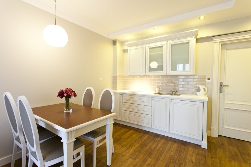 Compact suite kitchenette with white cabinets, subway-tile backsplash, sink, kettle and a four-seat wooden dining table with fresh flowers