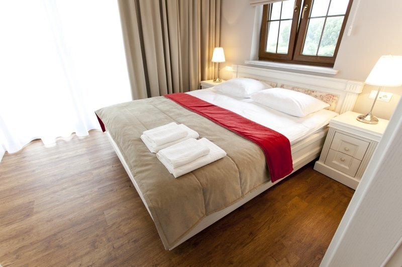 Bright double room with queen bed, beige duvet, red runner, folded towels on bed, bedside lamps, wooden floor and window view