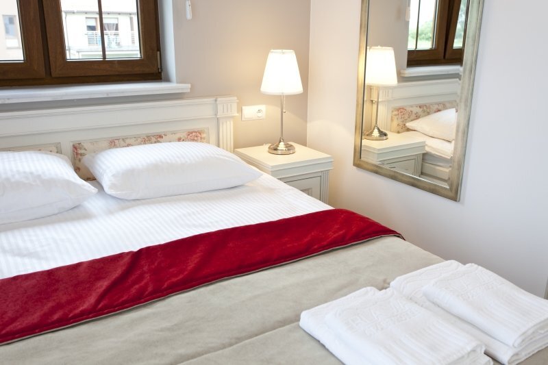 Bright double hotel room with white linens, red bed runner, bedside lamp, wall mirror and folded towels.