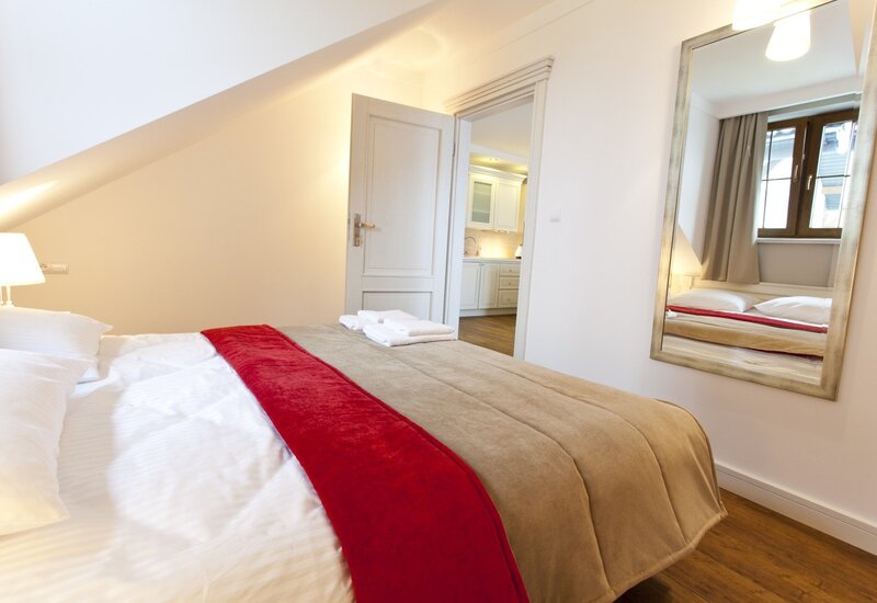 Cozy attic double room with sloped ceiling, double bed with white linens, beige bedspread and red runner, towels and kitchenette view.