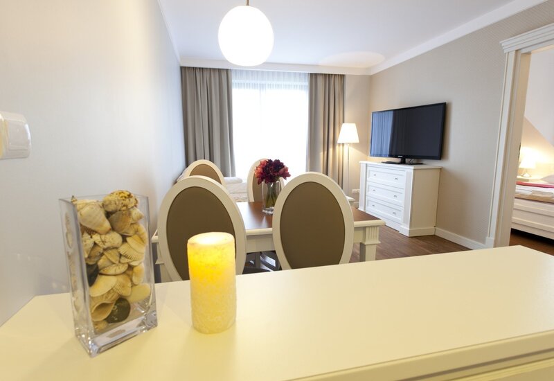 Bright modern hotel suite with dining table and oval-back chairs, vase of red flowers, TV on white dresser and visible bedroom.