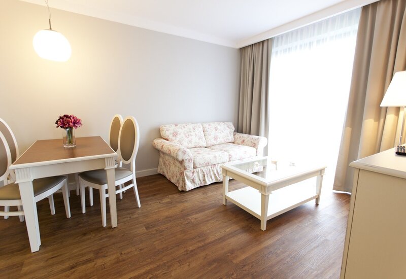 Bright hotel living area with floral sofa, white coffee table, dining table with chairs, hardwood floor and sheer curtains