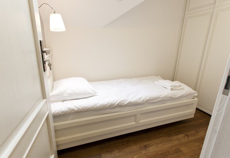Cozy single attic room with white single bed, crisp linens, folded towels, bedside lamp and built-in wardrobe over wooden floor