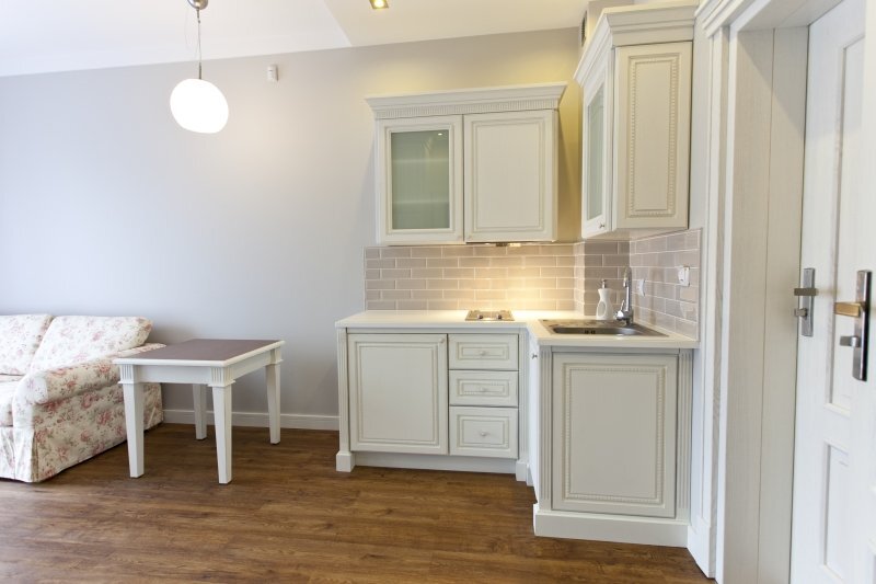 Studio suite kitchenette with cream paneled cabinets, subway-tile backsplash, two-burner stove, sink, floral sofa and dining table