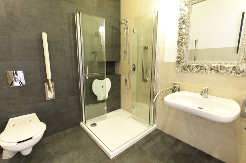 Accessible hotel bathroom with glass walk-in shower, fold-down seat, wall-mounted toilet, grab bars, decorative mirror.