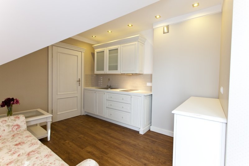 Cozy attic studio with white kitchenette, sink and hob, warm wood floor, recessed ceiling lights, floral sofa and side table with vase.