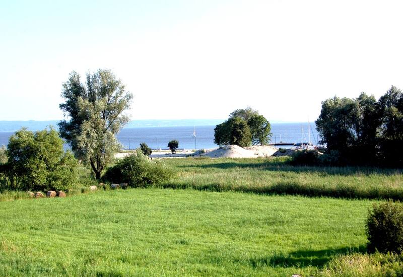 Green meadow and trees leading to a sandy beach and small marina with sailboats on a calm sea under clear sky
