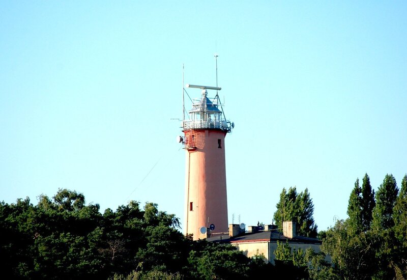 Peach-colored lighthouse with glass lantern and radar mast rising above pine trees, attached service building with rooftop antennas