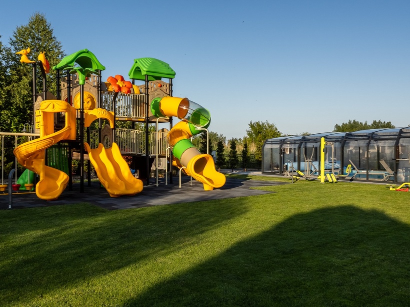 Plac zabaw dla rodzin z żółtymi spiralnymi i rurowymi zjeżdżalniami, zielone daszki nad trawnikiem obok zadaszonego basenu i siłowni