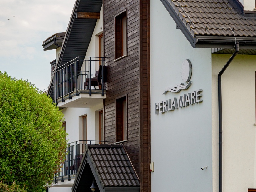Fasada hotelu w stylu chalet z drewnianą elewacją, dachówką, balkonami, przytulnym wejściem i napisem 'PERLA MARE', otoczona zielenią