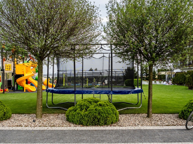 Zielony trawnik hotelowy z ogrodzonym trampolinem, kolorowym placem zabaw, rowerami przy ścieżce i gośćmi piknikującymi przy tarasie.