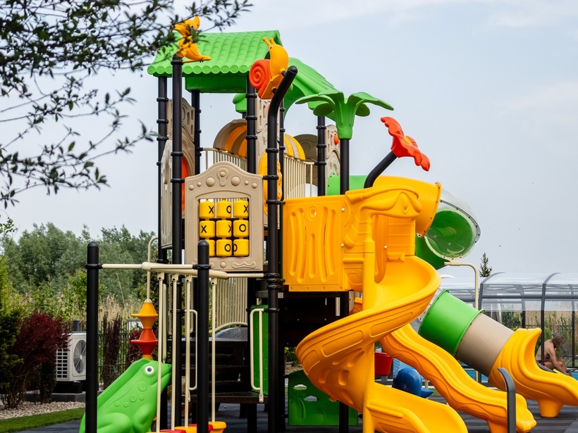 Kolorowy zewnętrzny plac zabaw dla dzieci z zielonym daszkiem, żółtą spiralną i tubową zjeżdżalnią, panelem kółko‑krzyżyk i sztuczną trawą.