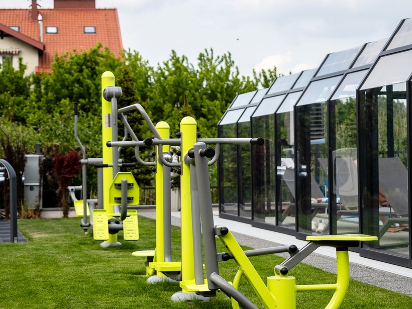 Outdoor fitness stations on a manicured lawn beside a glass-enclosed pool with sun loungers and garden views.