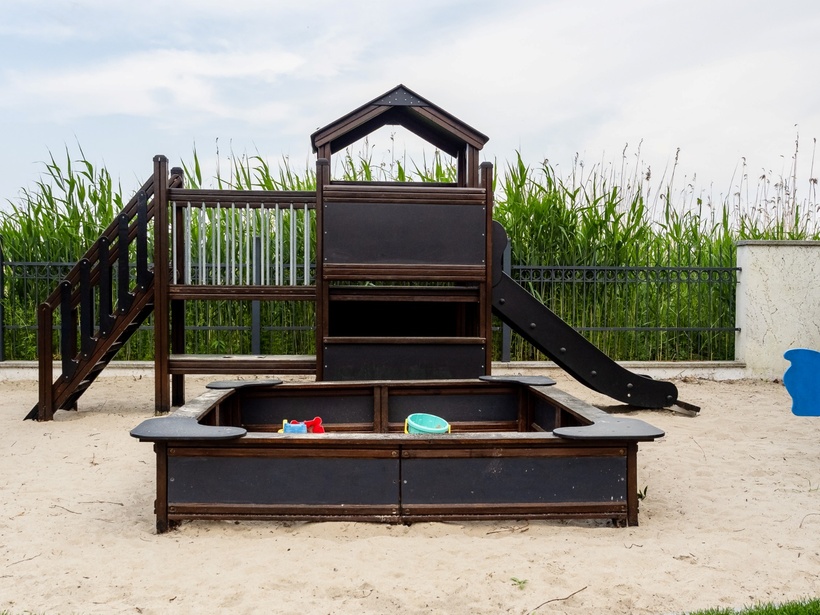 Piaszczysty plac zabaw dla dzieci z drewnianym domkiem i zjeżdżalnią, piaskownicą z zabawkami i dwoma niebieskimi bujakami.