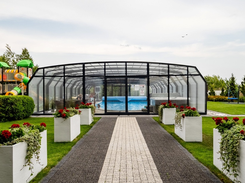 Covered glass-enclosed pool with lounge seating, central paved path lined by white planters with red flowers and a nearby children's playground