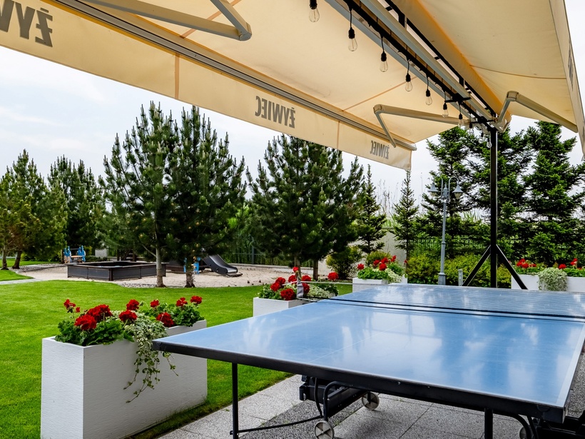 Stolik do ping-ponga na zewnątrz pod dużą markizą, zadbany trawnik, czerwone donice z kwiatami i plac zabaw wśród sosen.