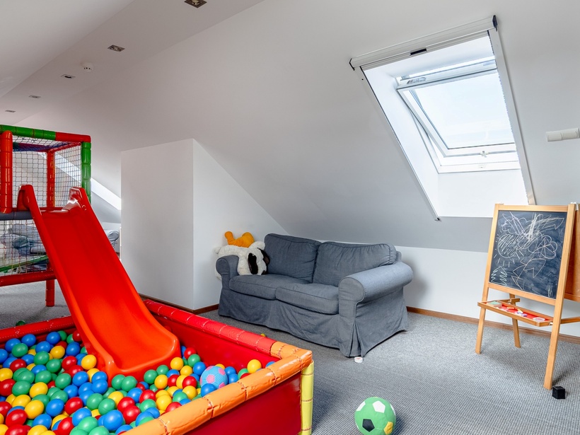 Hotelowy pokój zabaw z kolorowym basenem kulkowym i czerwoną zjeżdżalnią, szarą sofą pod oknem dachowym i kredową tablicą.