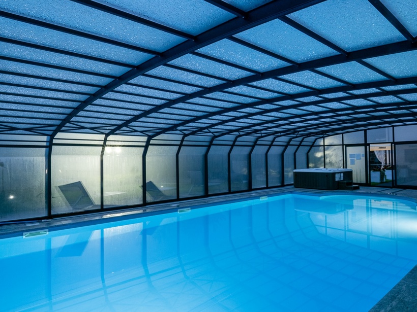 Heated indoor pool with blue ambient lighting, arched glass enclosure, lounge chairs and a hot tub for relaxed spa-style evenings