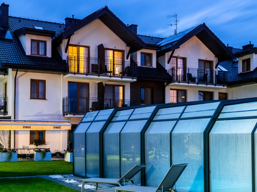 Alpine-style three-story hotel with warm-lit balconies, covered glass pool enclosure, lawn and poolside loungers at dusk.