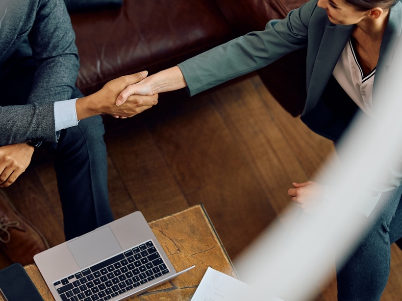 Geschäftlicher Handschlag in der Hotel-Lounge auf Ledersofa, hölzerner Couchtisch mit Laptop und Unterlagen