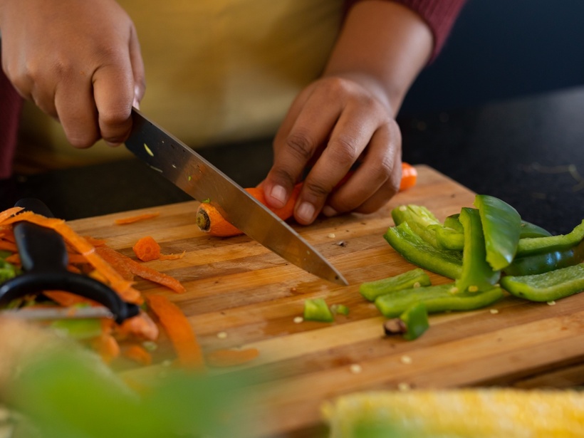 Gast schneidet Karotten auf einem Holzbrett mit Messer, grüne Paprikastreifen und Sparschäler daneben