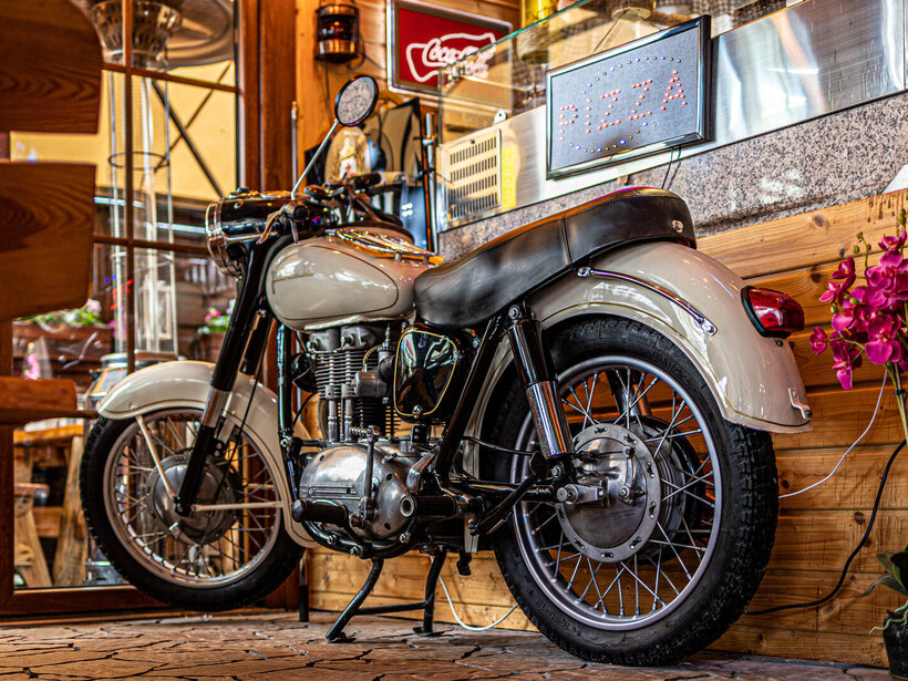 Vintage-Motorrad neben hölzerner Pizza-Theke mit leuchtendem 'Pizza'-Schild, Orchideen und gemütlichem, rustikalem Café-Ambiente.