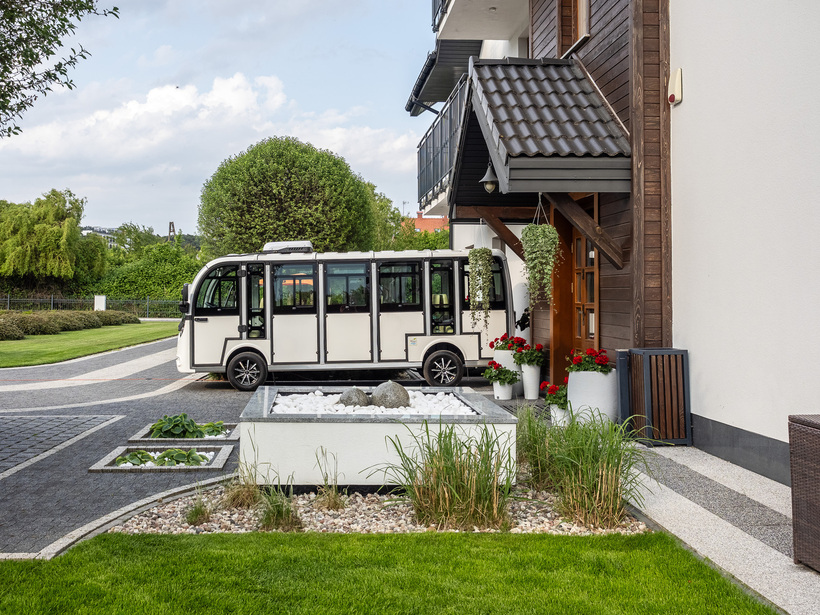 Shuttle hotelowy zaparkowany przy zadaszonym drewnianym wejściu, wiszące rośliny, czerwone pelargonie, kamienna dekoracja i zadbany trawnik