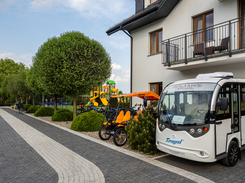 Fasada aparthotelu z busem transferowym, wypożyczalnią rowerów i pomarańczowym quadem rodzinnym, plac zabaw i przycięte drzewa