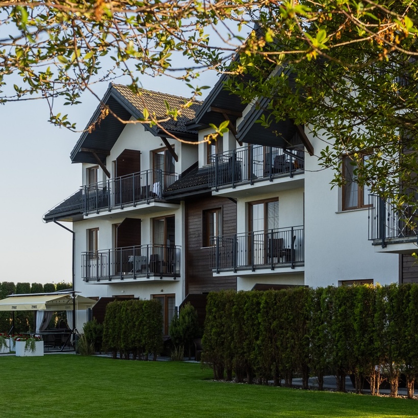 Trzypiętrowy hotel w stylu chalet z prywatnymi balkonami, drewnianymi akcentami, zadbaną trawą i zadaszonym tarasem jadalnym