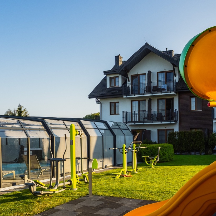 Nasłonecznione tereny hotelu rodzinnego z przeszklonym zadaszonym basenem, zewnętrzną siłownią na trawniku i pomarańczową zjeżdżalnią przy balkonach.