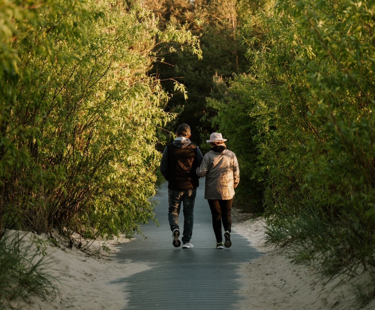 Couple walking arm-in-arm along a sandy boardwalk framed by lush dunes, tranquil beach-access path at golden hour