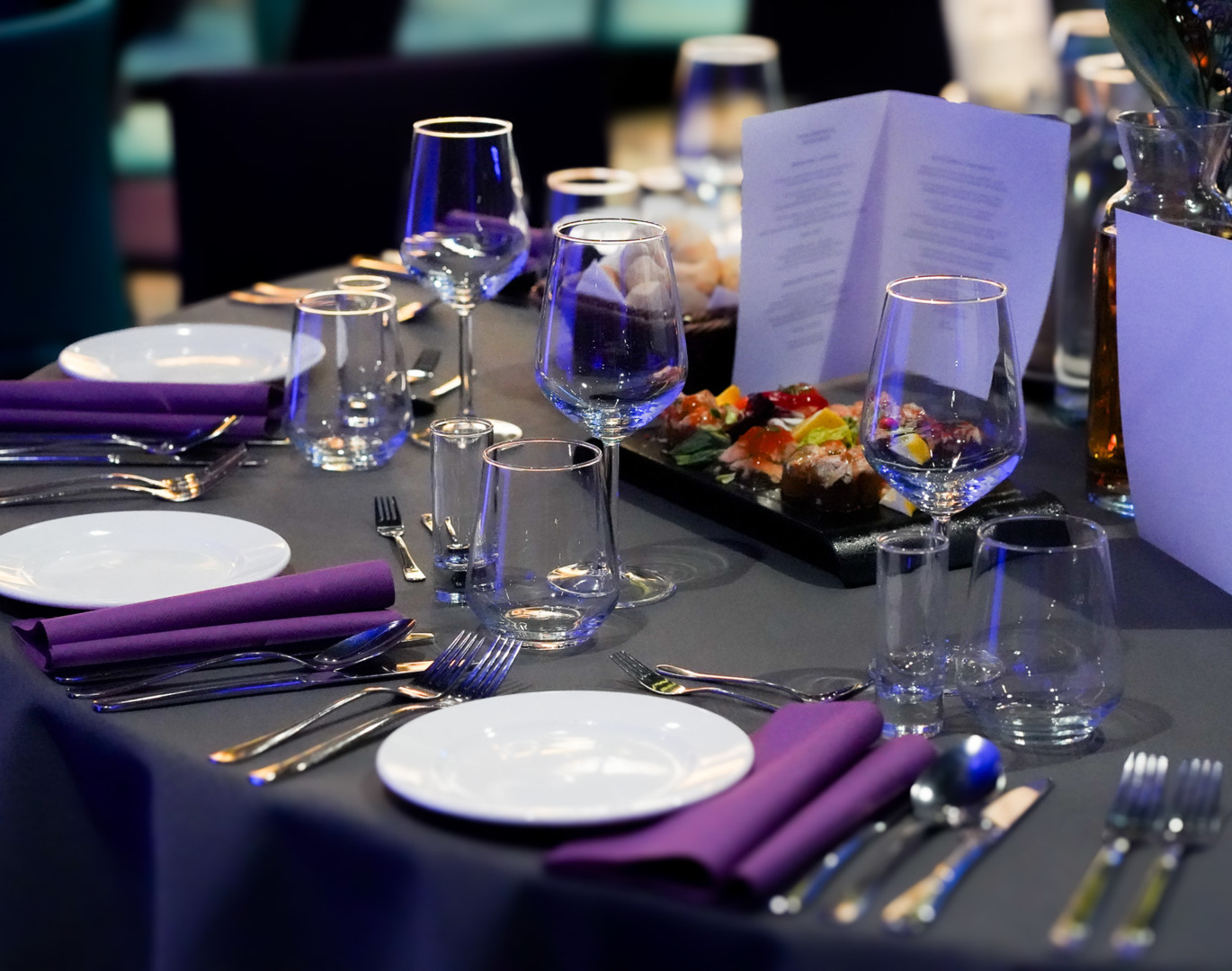 Hotel-Esstisch mit grauem Tischläufer, violetten Servietten, Weingläsern, Vorspeisenplatte und Menükarte