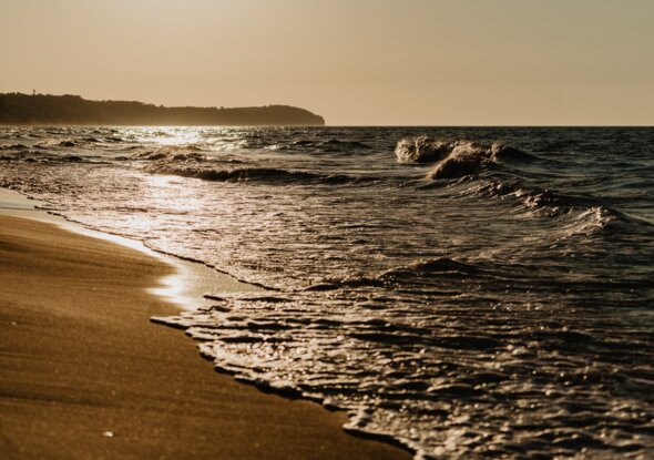 Goldener Sonnenuntergang über ruhigem Sandstrand, sanfte Wellen am Ufer und eine entfernte, bewaldete Landspitze