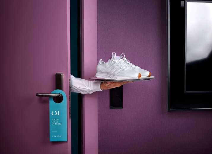 Hand eines Hotelmitarbeiters reicht saubere weiße Sneakers auf einer silbernen Platte durch eine violette Zimmertür mit Türanhänger ‚Please clean my room‘.