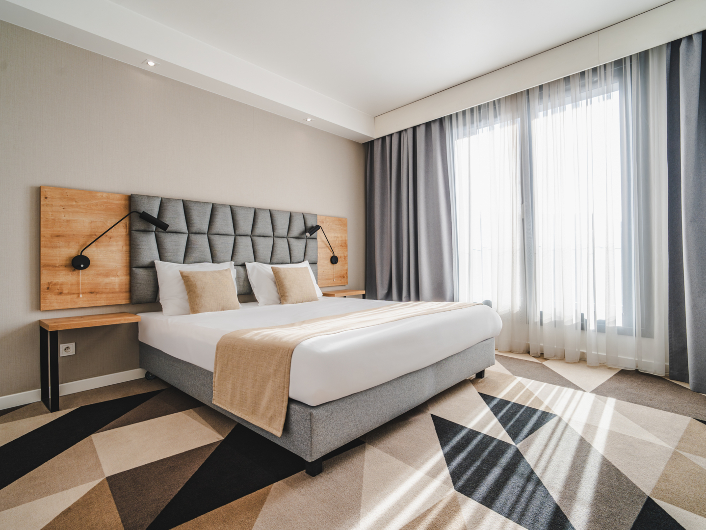 Helles, modernes Hotelzimmer mit großem Bett, grauem gepolstertem Kopfteil, Holzakzenten und Tageslicht durch transparente Vorhänge.