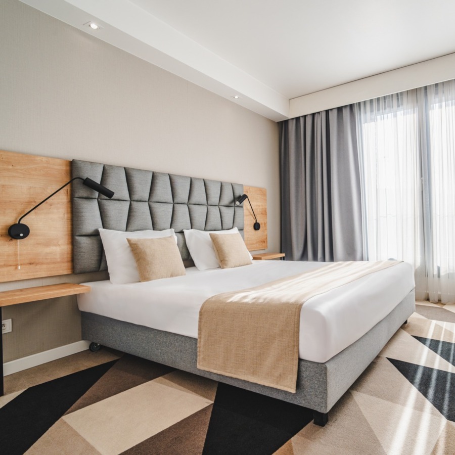 Moderner Hotelzimmer mit großem Bett, grauem gepolstertem Kopfteil und natürlichem Licht durch bodentiefe Fenster mit Vorhängen.