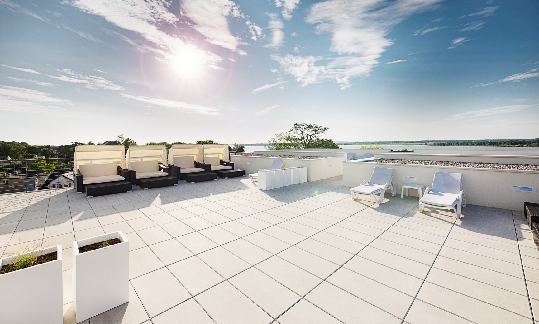 Dachterrasse mit schattigen Daybeds und Liegestühlen, Panoramablick aufs Wasser und sonniger Himmel
