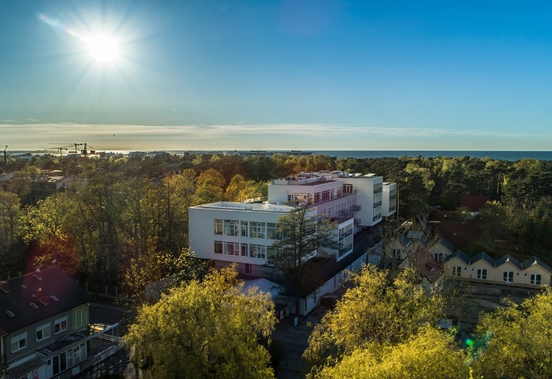 Nowoczesny biały hotel z tarasami na dachu, otoczony drzewami, ze słonecznym widokiem na linię brzegową i morze.