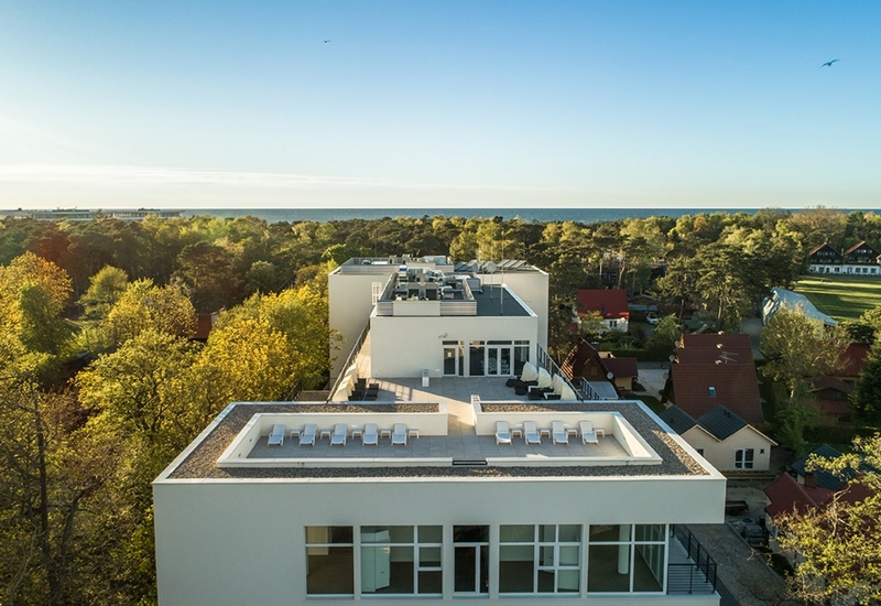 Taras na dachu z leżakami w nowoczesnym białym hotelu, otoczony słonecznym lasem nadmorskim i widokiem na morze.