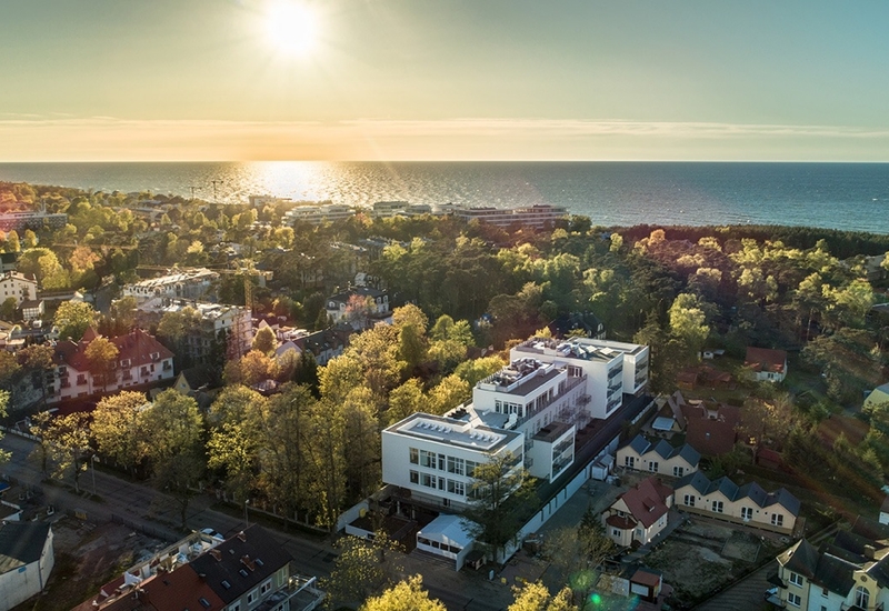 Nowoczesny biały hotel z tarasami i widokiem na morze, otoczony zielonymi drzewami i nadmorską promenadą o zachodzie słońca.